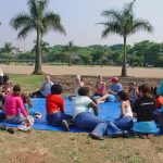 Grupo de pessoas com e sem deficiência estão reunidos em círculo sobre uma lona azul, em cima da grama. Eles conversam entre si. Está um dia ensolarado no parque e há coqueiros e árvores ao fundo.
