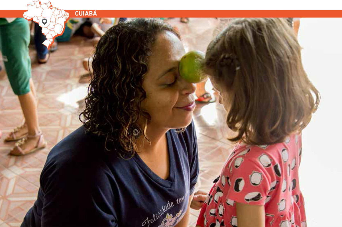 Uma de frente para a outra, educadora e menina usam as testas para equilibrar uma laranja entre elas. Para manter a mesma altura, a professora está agachada. Acima da imagem, mapa do Brasil com marcação que destaca cidade de Cuiabá.