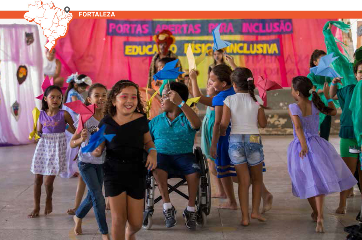Crianças correm e dançam em quadra escolar, usando pinturas no rosto e vestindo fantasias. Elas seguram pássaros feitos com papel dobrado. Um garoto em cadeira de rodas está no centro. Acima da imagem, mapa do Brasil com marcação que destaca cidade de Fortaleza.