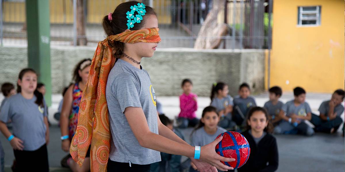 Menina de olhos vendados com um tecido segura bola com uma das mãos. Atrás, estudantes acompanham a atividade sentados na quadra.