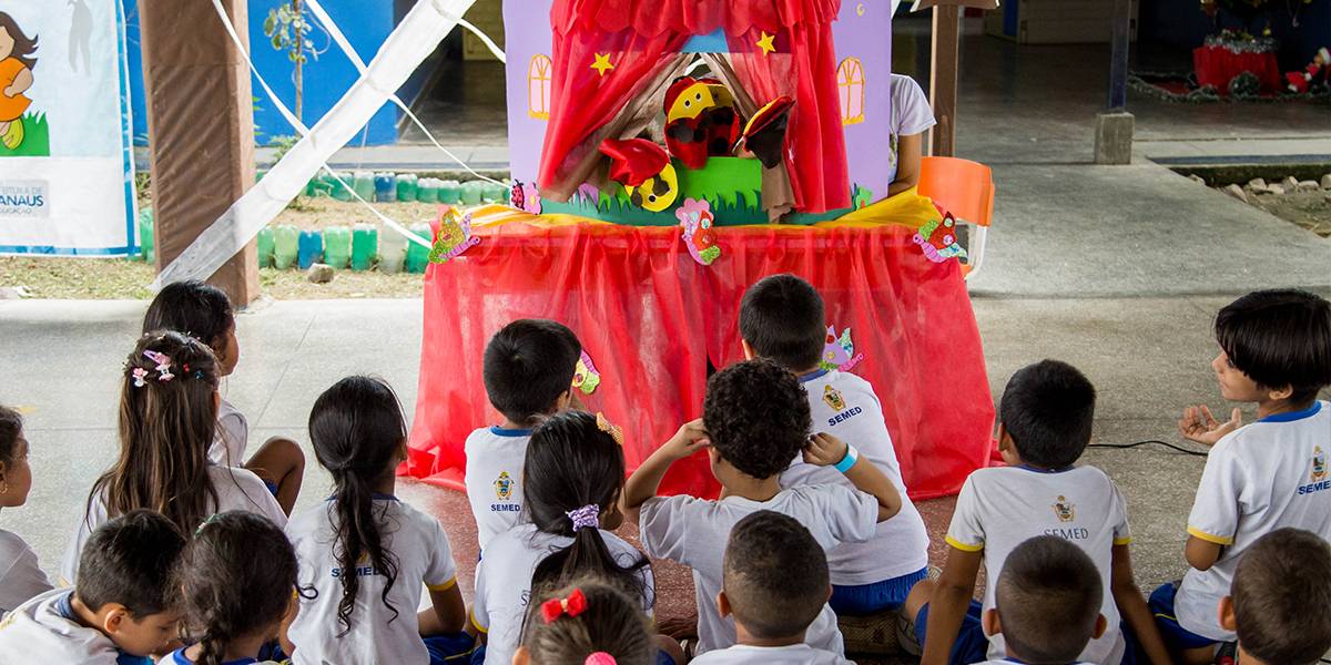 Crianças sentadas no pátio da escola assistem ao teatro de fantoches montado sobre uma mesa enfeitada com tecido vermelho.