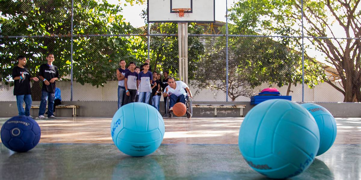 Homem em cadeira de rodas lança bola de basquete no chão em direção a um conjunto de bolas azuis adiante. Atrás dele, adolescentes observam.