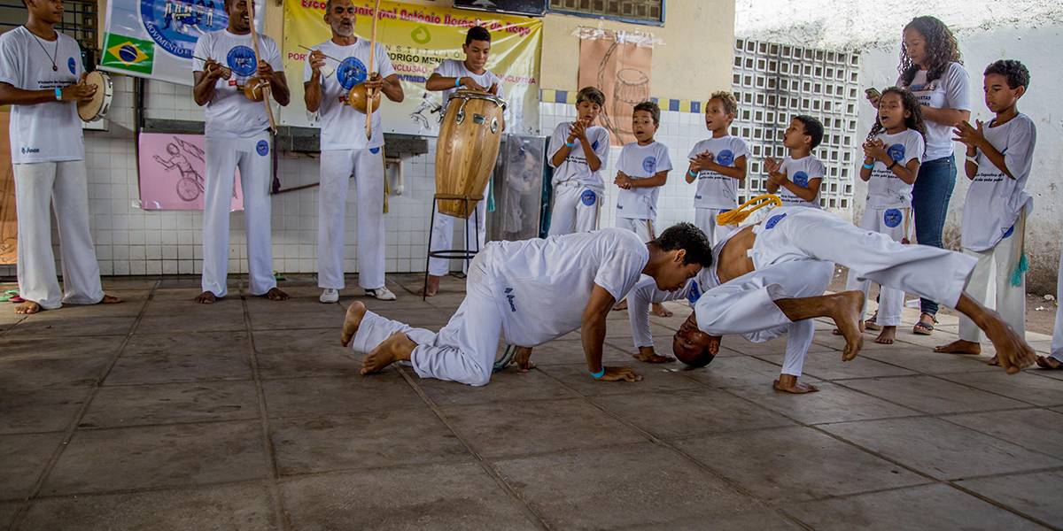 Professor e estudante com deficiência física jogam capoeira com movimentos no chão.