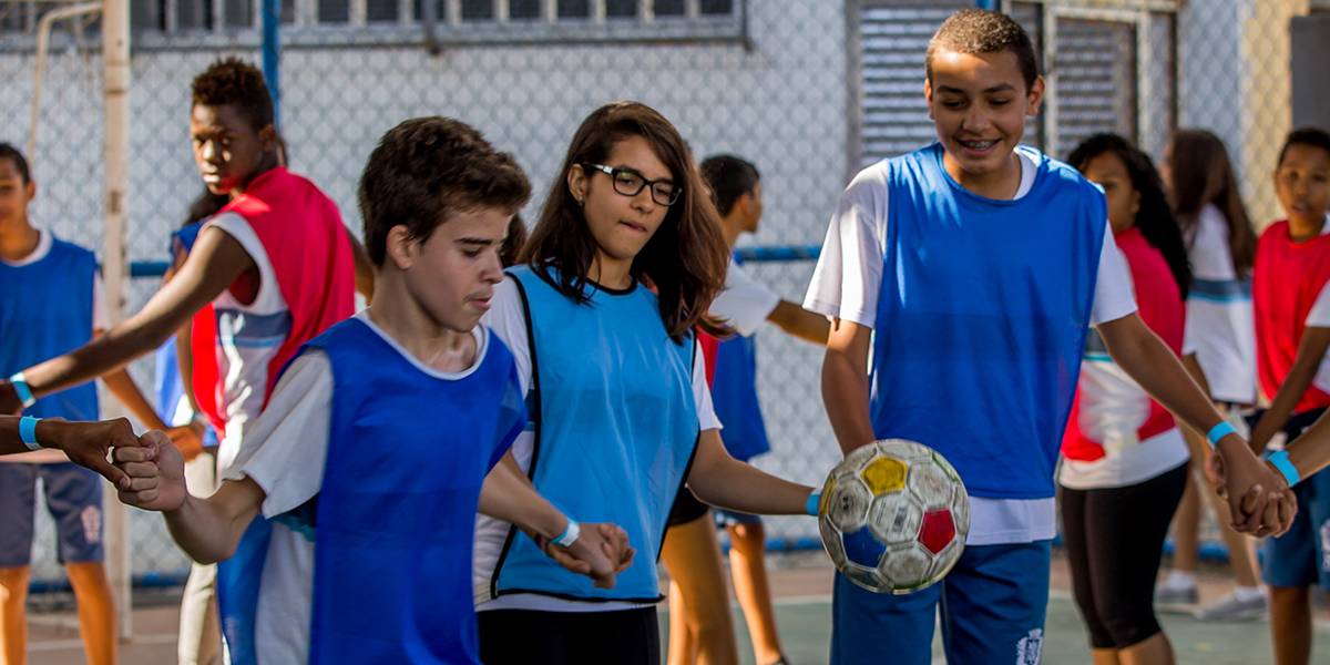 Garoto de mãos dadas com colega de time se prepara para chutar bola de futebol para frente. Os outros jogadores acompanham a jogada ao fundo.