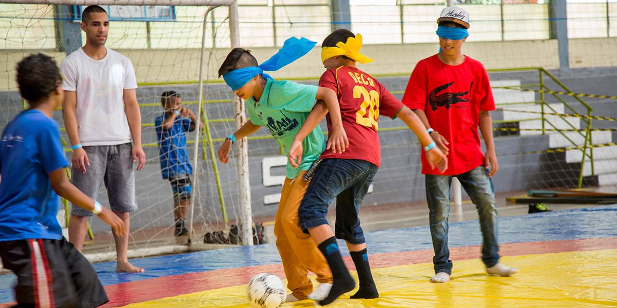 Dois garotos com os olhos vendados por tecidos disputam bola em jogo de futebol. Atrás, outro jogador vendado acompanha a jogada.