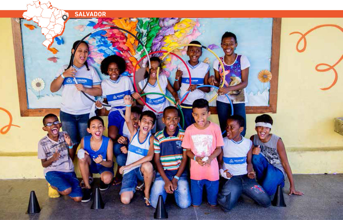 Crianças posam sorridentes segurando bambolês coloridos que formam arcos olímpicos. Acima da imagem, mapa do Brasil com marcação que destaca cidade de Salvador.