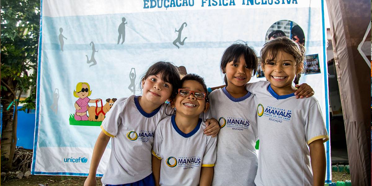 Quatro crianças enfileiradas sorriem. Elas estão com os braços entrelaçados sobre os ombros uma das outras. Ao fundo, cartaz com o logotipo da prefeitura de Manaus e os dizeres “Portas abertas para inclusão educação física inclusiva”.