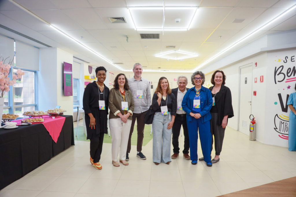 Foto dos sete jurados do Prêmio Educador Nota 10. Sete pessoas posam para a foto, sendo uma mulher negra, quatro mulheres brancas e dois homens brancos. Elas estão em pé e sorriem. À esquerda, uma mesa com salgados servidos. Fim da descrição. 