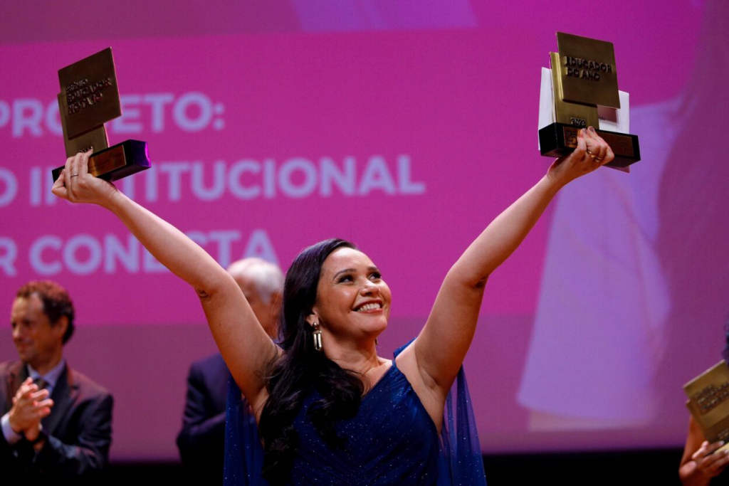 Foto da Educadora do Ano. Uma mulher parda sorridente com os braços erguidos exibe dois troféus, um em cada mão. Ela tem os cabelos negros e compridos e usa um vestido azul. Fim da descrição.  