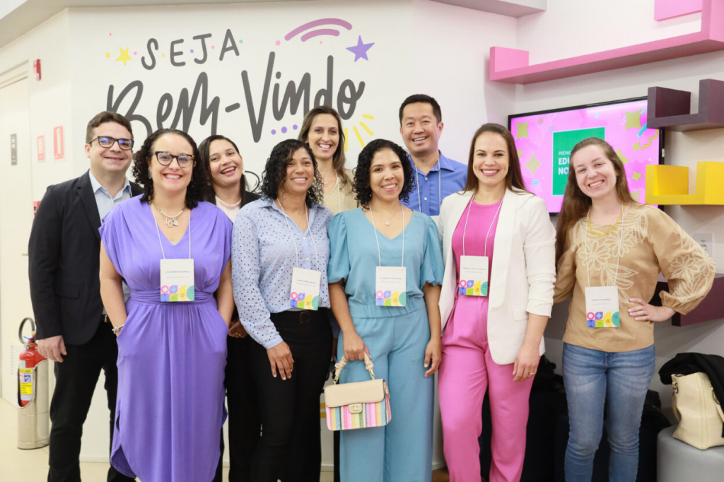 Foto dos nove educadores vencedores do Prêmio Educador Nota 10 de 2023. Um grupo formado por nove pessoas, sendo um homem asiático, um homem branco, duas mulheres negras com pele de tom claro, uma mulher parda, e quatro mulheres brancas. As pessoas vestem roupas em tons de azul, rosa, roxo e bege. Todos estão em pé e sorriem para a foto. Fim da descrição. 